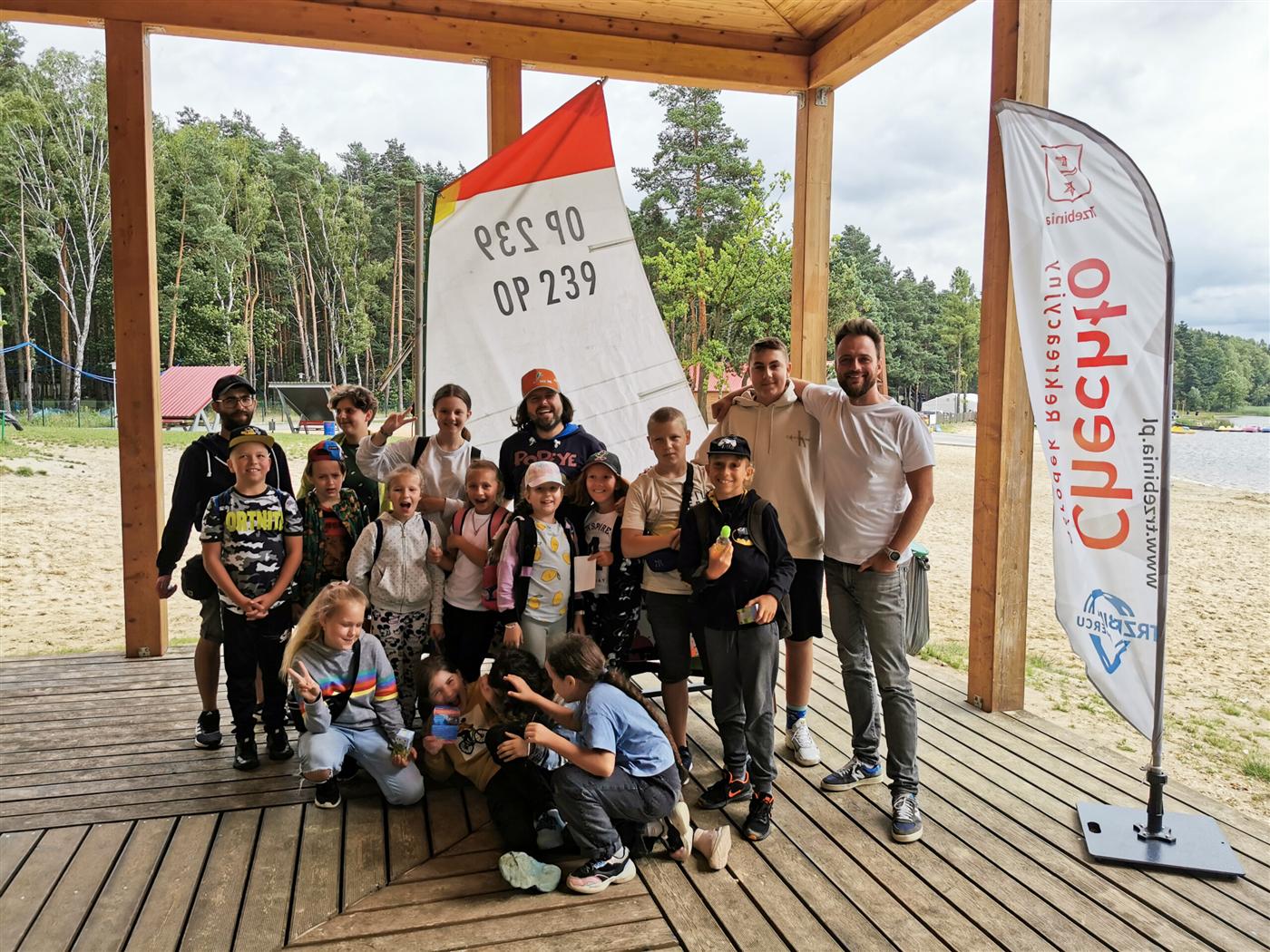 Grupa dzieci i kadry Akademii Piratów na pomoście nad Chechłem, żagiel Optimista w tle.