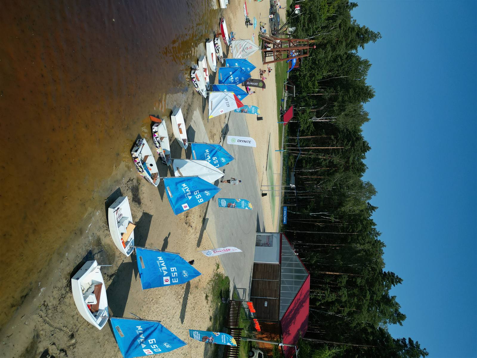 Łódki klasy Optimist z niebieskimi żaglami na plaży Chechła, flaga OXYNITE.